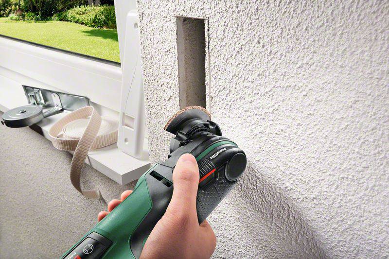 Een hand houdt een multitool vast dat een rechthoekig gat in een muur snijdt, vermoedelijk voor de installatie van een schakelaar of stopcontact.