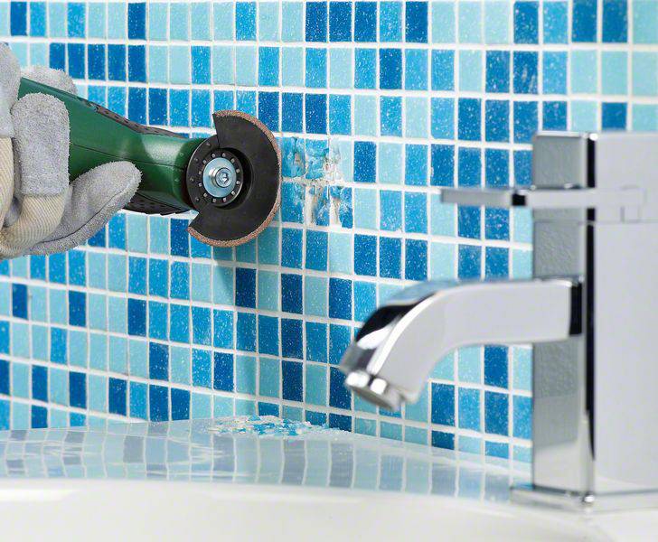 Een persoon gebruikt een elektrisch gereedschap om blauwe mozaïektegels aan een muur te snijden. Op de voorgrond is een waterkraan zichtbaar.