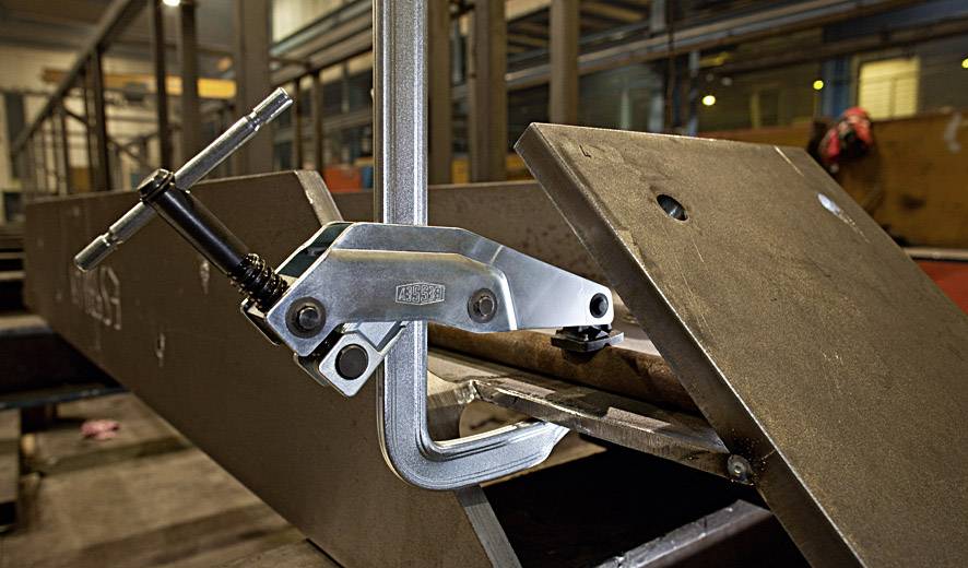 Industriehal met metaalwerken: Een C-klem bevestigt een metalen onderdeel aan een stalen ligger. Machines en gereedschappen op de achtergrond.