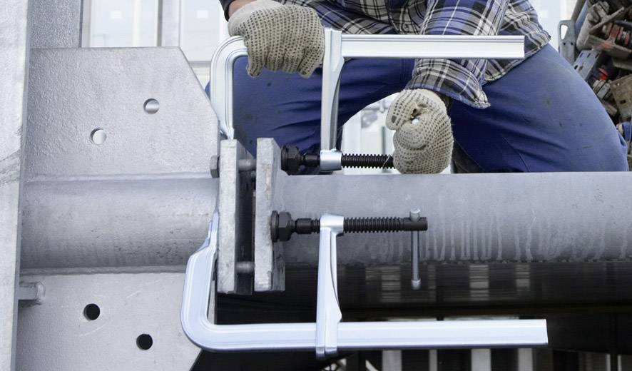 Een persoon in werkkleding bevestigt een metalen buis aan een constructie met behulp van klemmen. Grote handschoenen beschermen de handen.