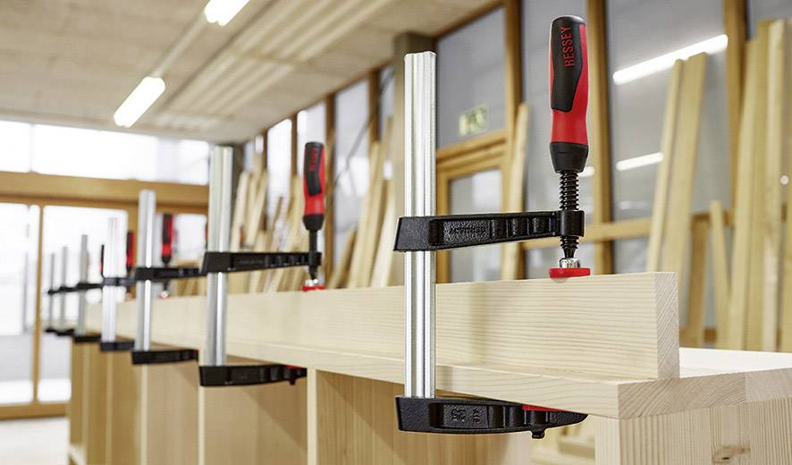 Verschillende houten planken worden met lijmklemmen op werkbanken in een werkplaats vastgezet. Op de achtergrond zijn ramen en houten frames zichtbaar.