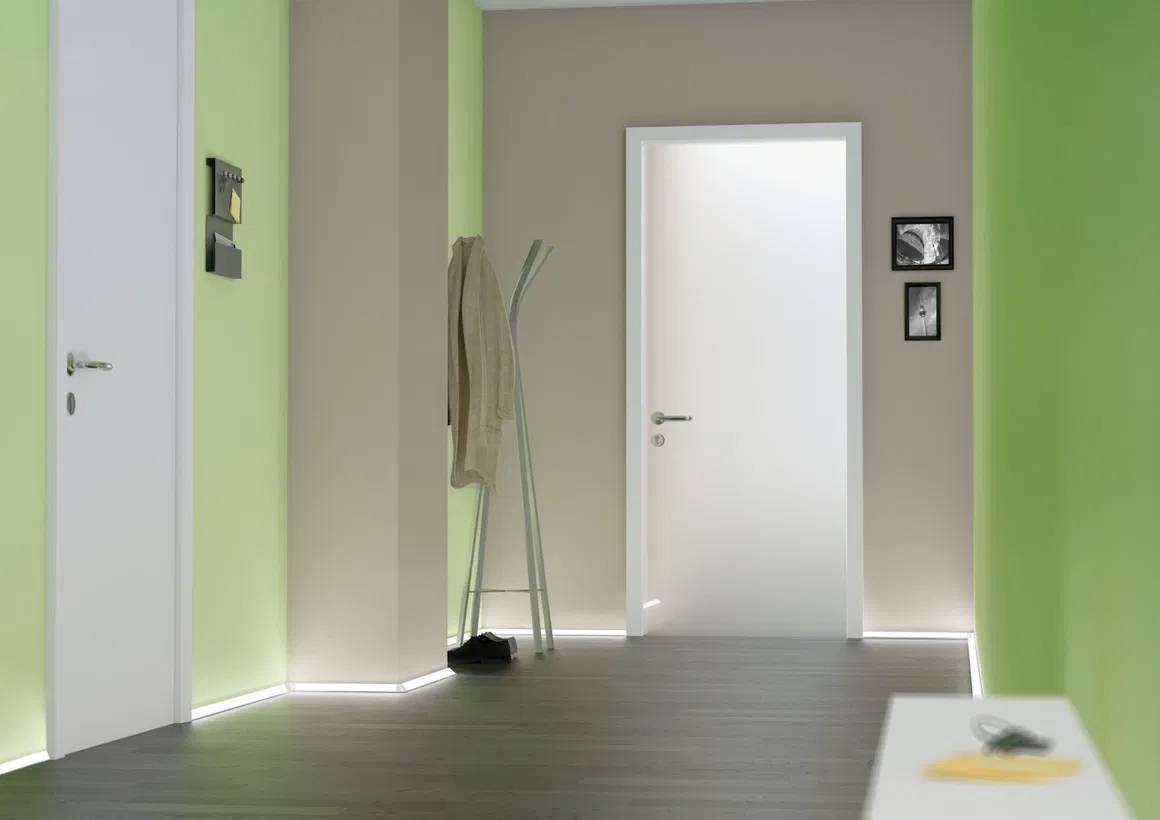 Een moderne hal met groen en beige geschilderde muren, donkere houten vloer en een witte deur, met een kapstok waaraan een beige jas hangt.