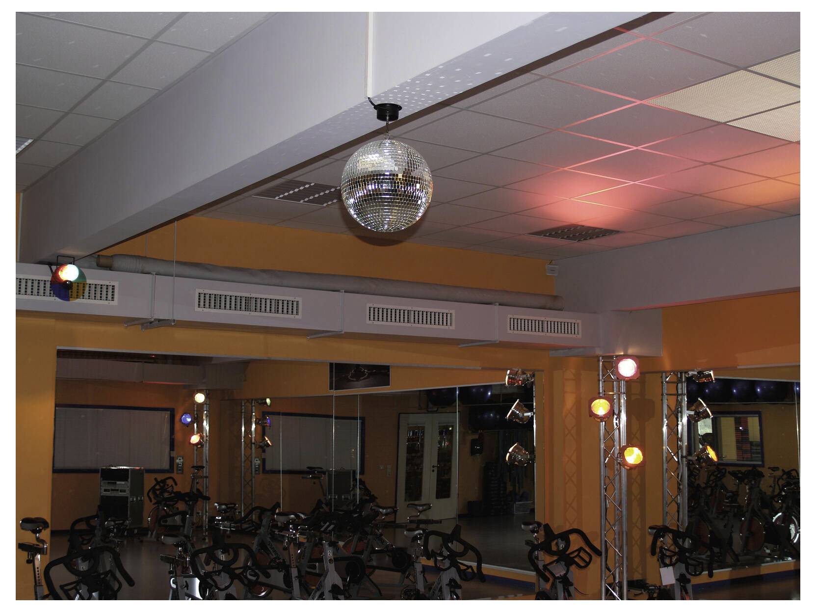 Een spiegelde discobol hangt in een gymnastiekzaal of studio met hometrainers, verlicht door gekleurde podiumlampen die reflecteren op het plafond en de muren.