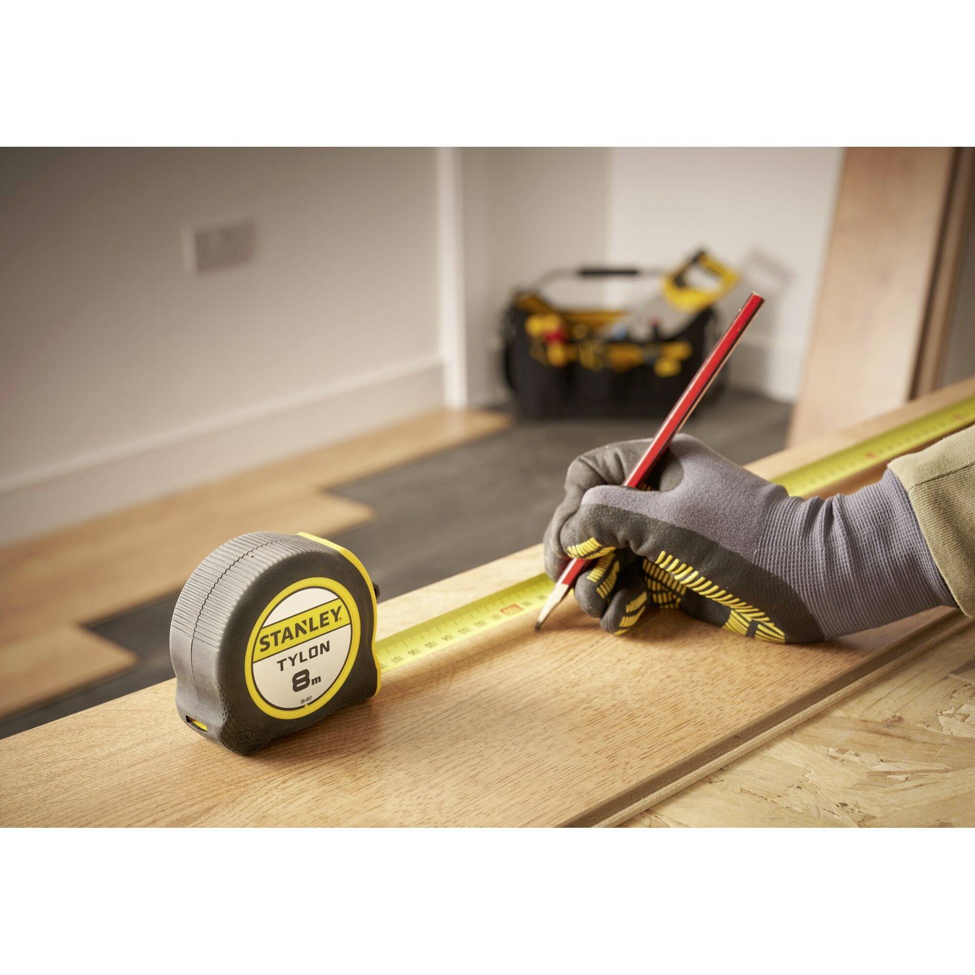 Een hand houdt een rode potlood vast, markeert een houten plank terwijl een meetlint de lengte opmeet. Op de achtergrond gereedschappen.