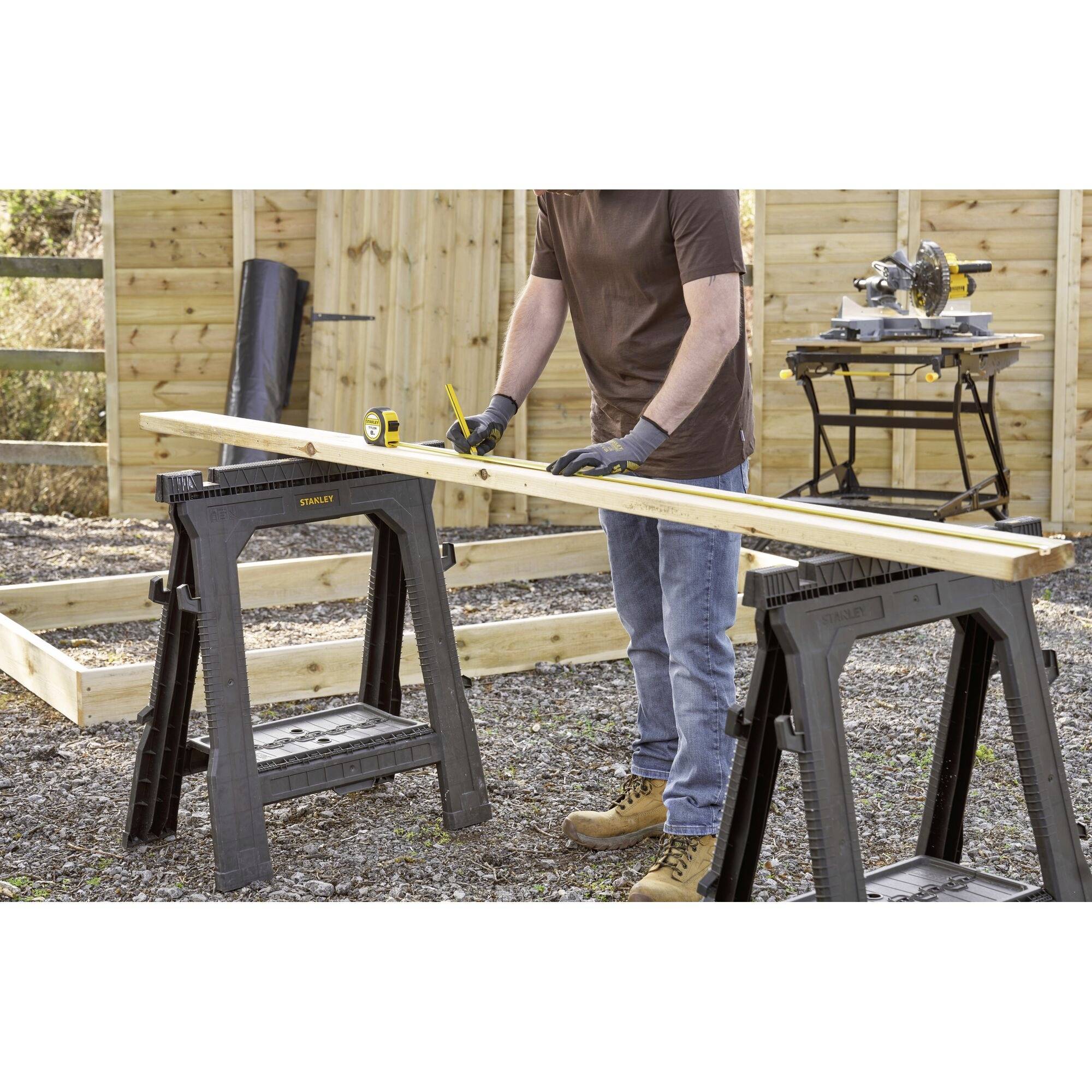 Een persoon meet een houten plank met een meetlint. Op de achtergrond zijn een zaag op een tafel en houten latten te zien.