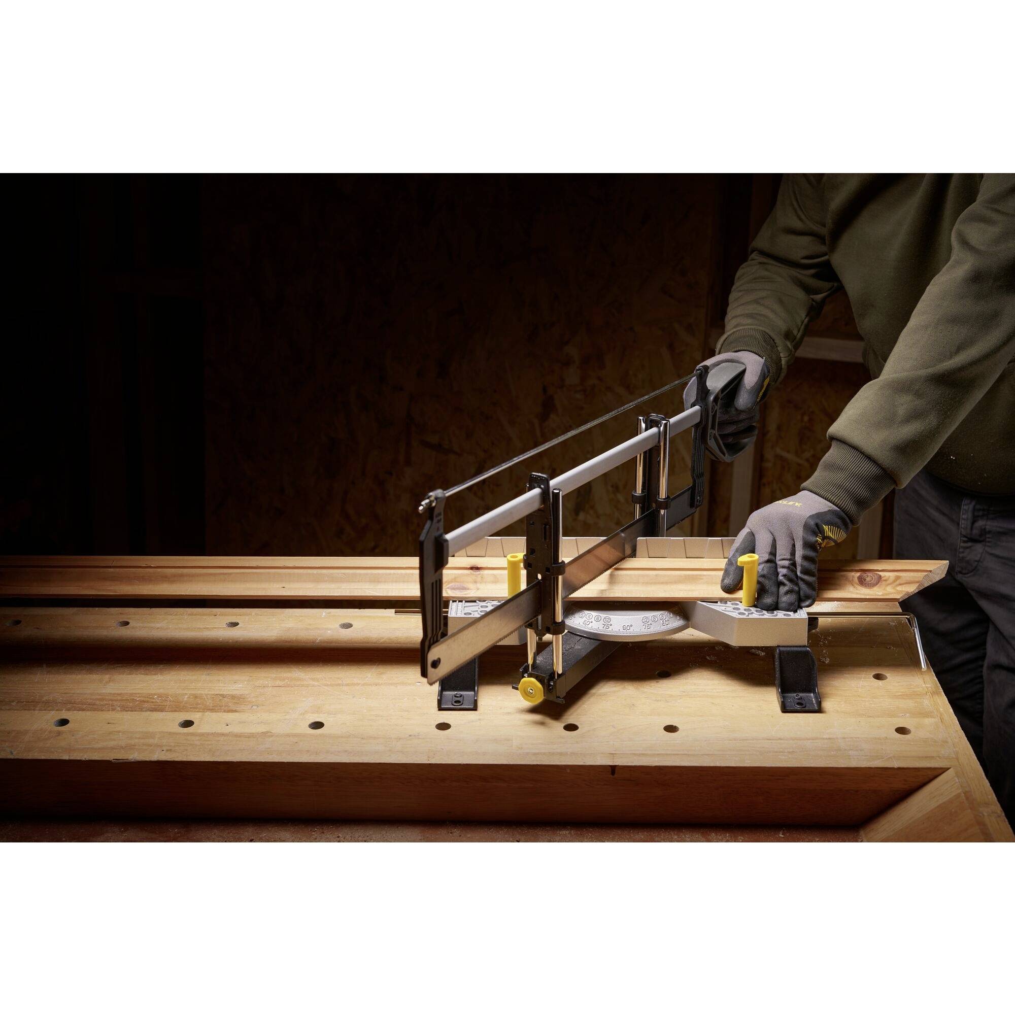 Een persoon bedient een verstekzaag aan een houten tafel. Hij draagt werkhandschoenen. De omgeving heeft een ambachtelijke uitstraling.