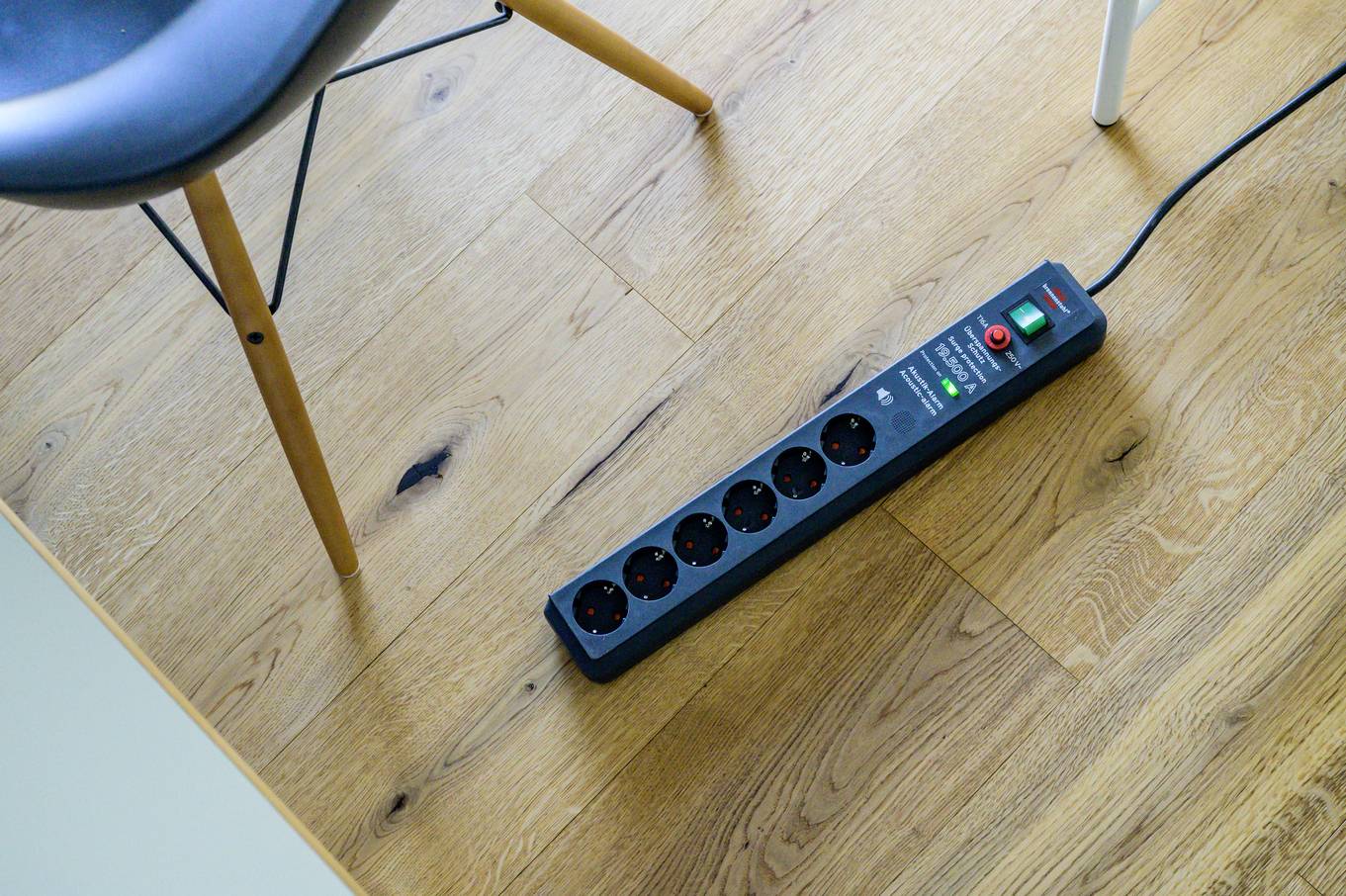 Een overspanningsbeveiligde stekkerdoos ligt op een houten vloer naast een stoel, klaar om meerdere apparaten aan te sluiten.