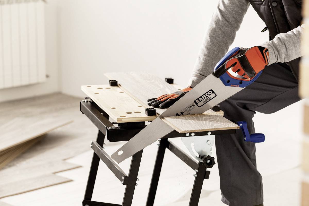 Een persoon zaagt een houten plank op een werkbank. Hij draagt werkhandschoenen en gebruikt een handzaag.
