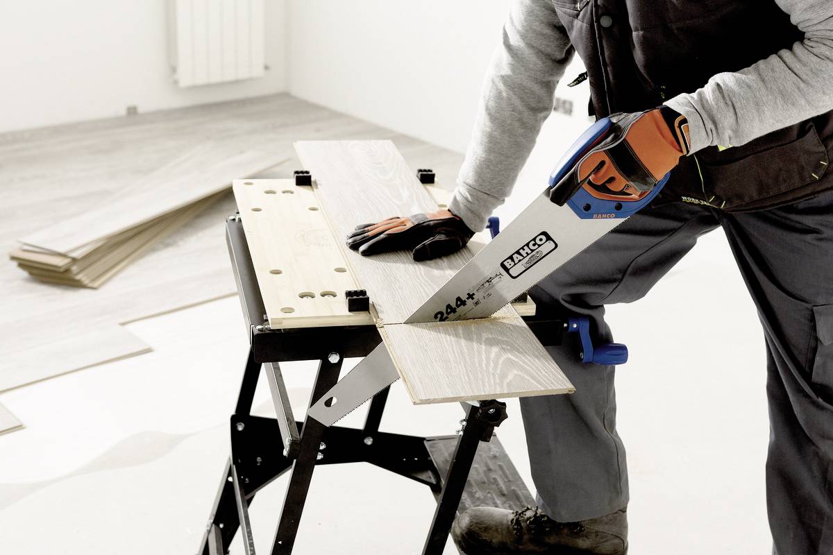 Een persoon zaagt een houten plank op een werktafel met een handzaag in een ruimte. Op de vloer liggen nog meer houten platen.