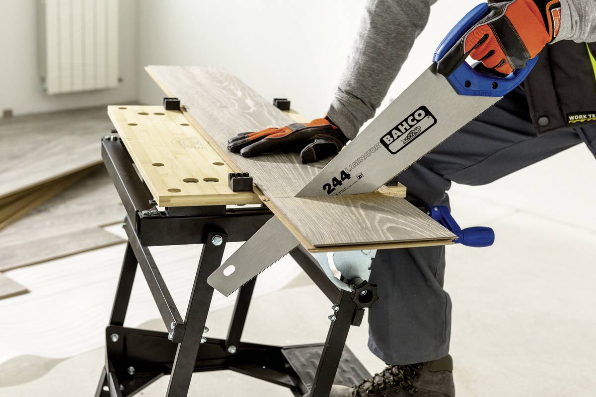 Een persoon zaagt met een handzaag een houten plank op een werkbank. Veiligheidskleding en handschoenen worden gedragen.