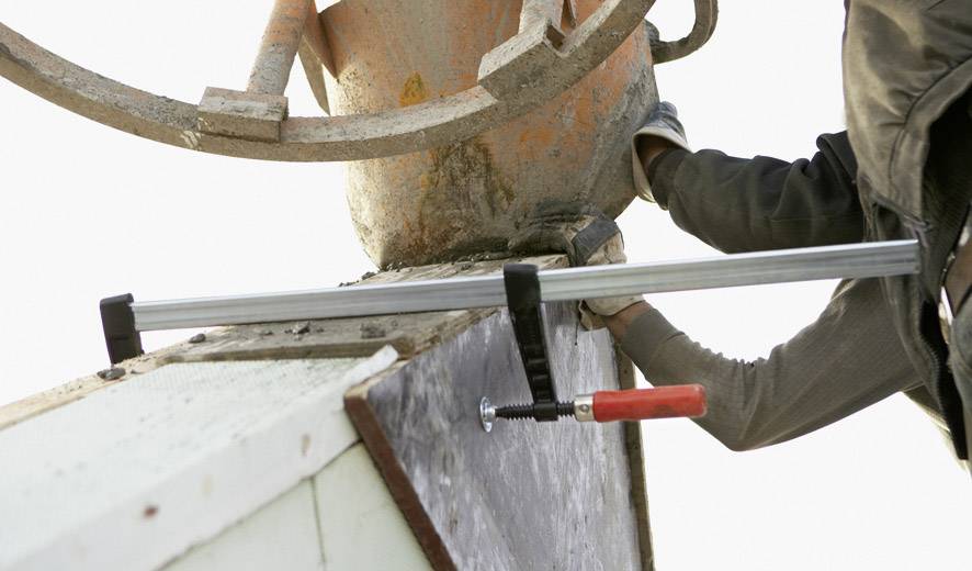 Een arbeider bevestigt een onderdeel met een klemhaak aan een metalen plaat. De omgeving doet denken aan een bouwplaats.