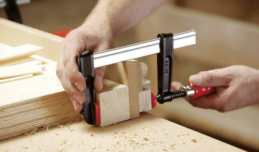 Een persoon klemt houten stukken met een klem vast op een werkbank. Houtspaanders liggen verspreid op het houtoppervlak.