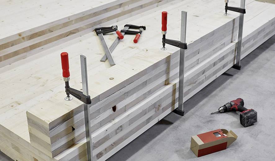 Stapel houten panelen die zijn vastgezet met rode klemmen. Naast de stapel liggen een boormachine en een lege verpakking op de vloer.