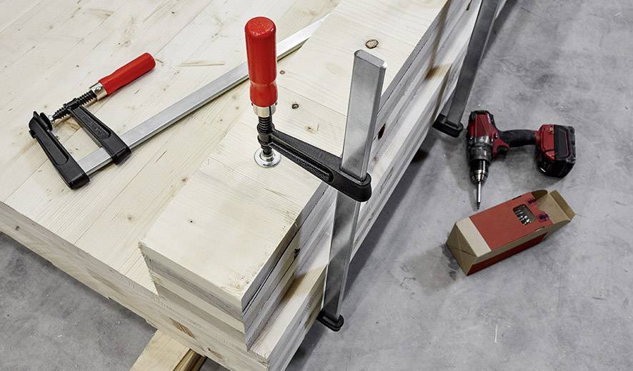 Houten planken worden met klemmen op een werkbank vastgezet. Ernaast liggen een zaag, een accuschroefmachine en een pak schroeven.