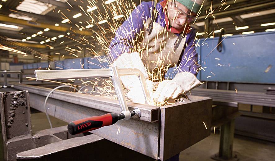 Een persoon in beschermende kleding last metaal in een fabriekshall. Vonken spatten rond. Op de achtergrond zijn gereedschappen en machines zichtbaar.