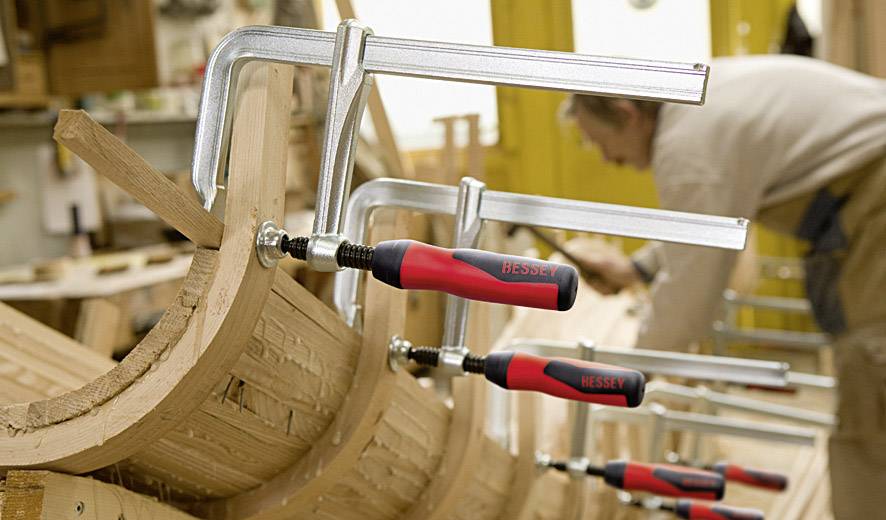 Een meubelmaker werkt aan een houten voorwerp dat met lijmklemmen is vastgezet. De werkplaats is op de achtergrond zichtbaar.