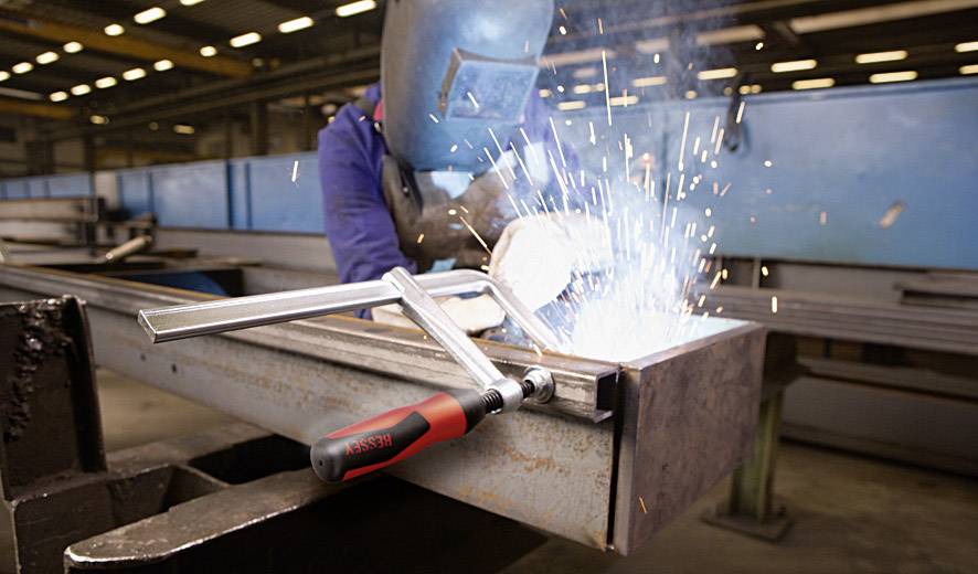 Een persoon last in een fabriekshal. Vonken spatten van het metalen stuk. Beschermende kleding en lasmasker worden gedragen.