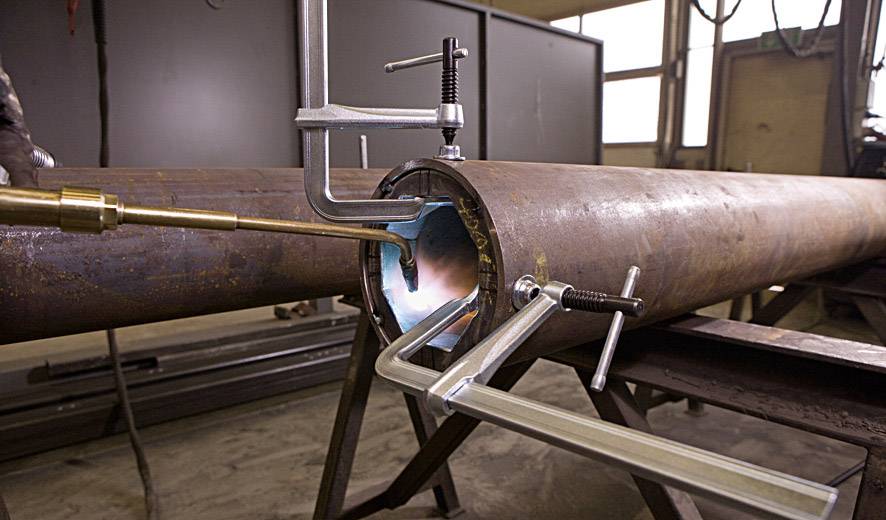 Een metalen buis wordt vastgeklemd terwijl er van binnenuit wordt gelast. Een vlam is zichtbaar in het binnenste. De omgeving is industrieel.