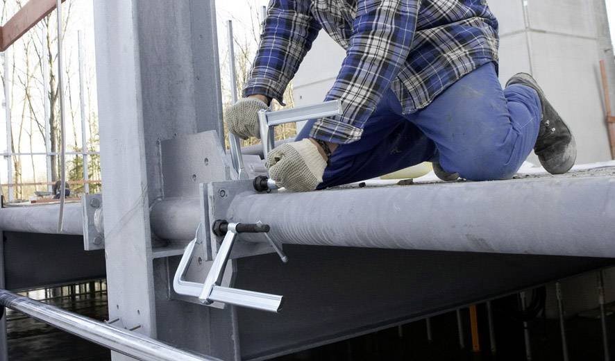 'Arbeider in beschermende uitrusting bevestigt metalen onderdelen op een bouwplaats, gebruikt klemmen om vast te zetten. Industriële omgeving.'