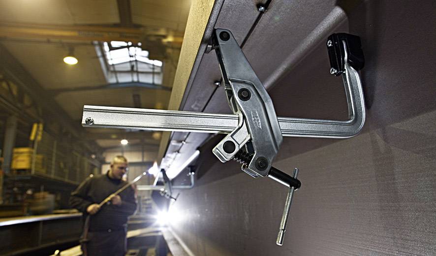 Een industriële scène met een grote metalen klem op de voorgrond, bevestigd aan een machine. Op de achtergrond werkt een onscherpe persoon.