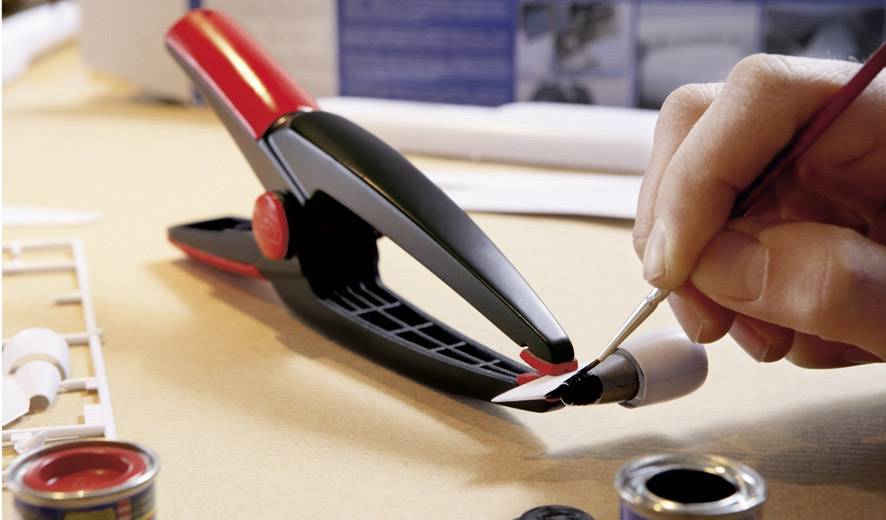 Een hand schildert met een penseel een vleugeldeel van een modelvliegtuig rood. Naast het onderdeel ligt een zwarte modelbouw-klem.