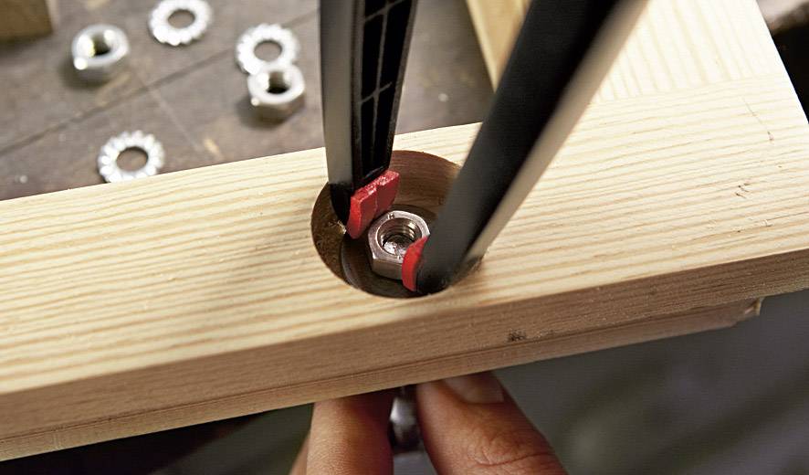 Een tang trekt een schroef uit een houten plank. De achtergrond toont een werkgebied met nog meer metalen moeren.