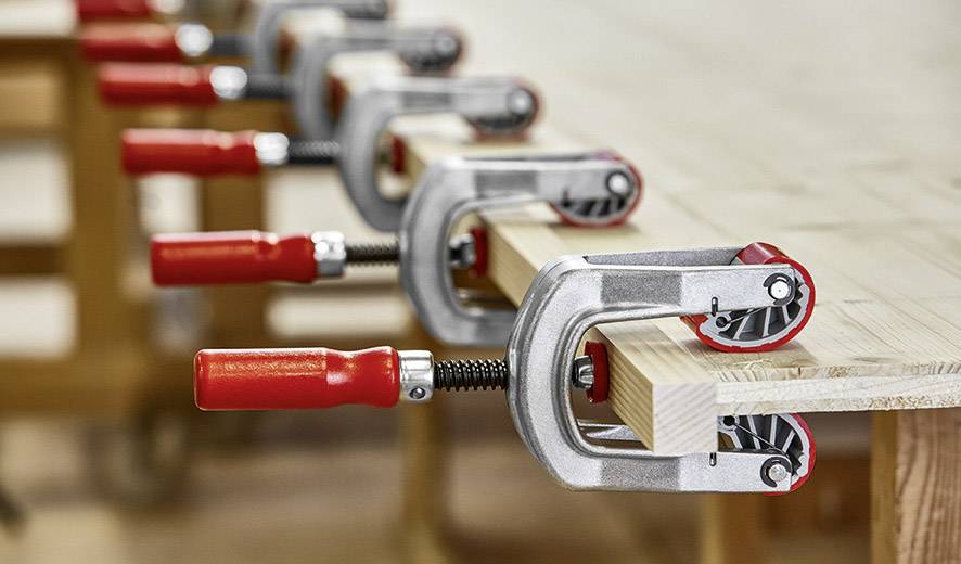 Hout wordt stevig vastgeklemd op een werkbank met behulp van meerdere lijmklemmen. De klemmen hebben rode handgrepen en houden het hout stabiel.