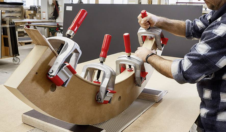 Een ambachtsman bewerkt een gebogen houten stuk dat met verschillende klemmen op een tafel is vastgezet, in een werkplaats.