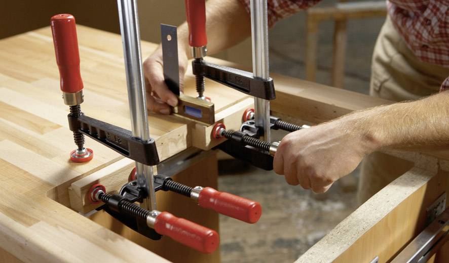 Twee handen die hout vastzetten met meerdere lijmklemmen op een werkbank. Een waterpas controleert de uitlijning.