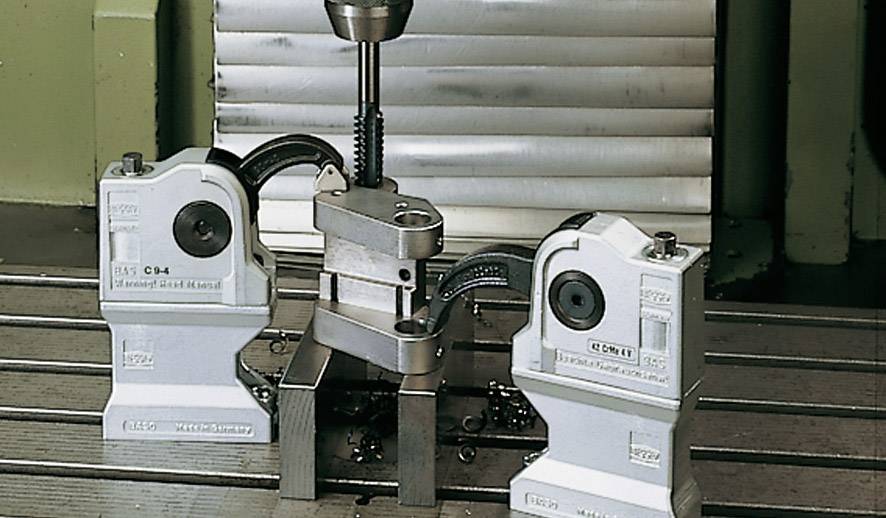 Een machine in een werkplaats die een werkstuk met twee klemmen vasthoudt. Een boor is boven het werkstuk gepositioneerd.