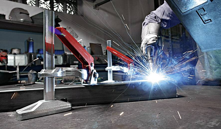 Een lasser werkt in een werkplaats, vonken vliegen rond. Metalen onderdelen zijn vastgeklemd, beschermende werkkleding wordt gedragen.