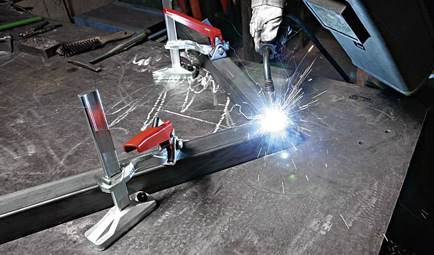 Een persoon last metalen onderdelen op een tafel, omringd door klemmen en vonkenslag. Beschermende kleding wordt gedragen.