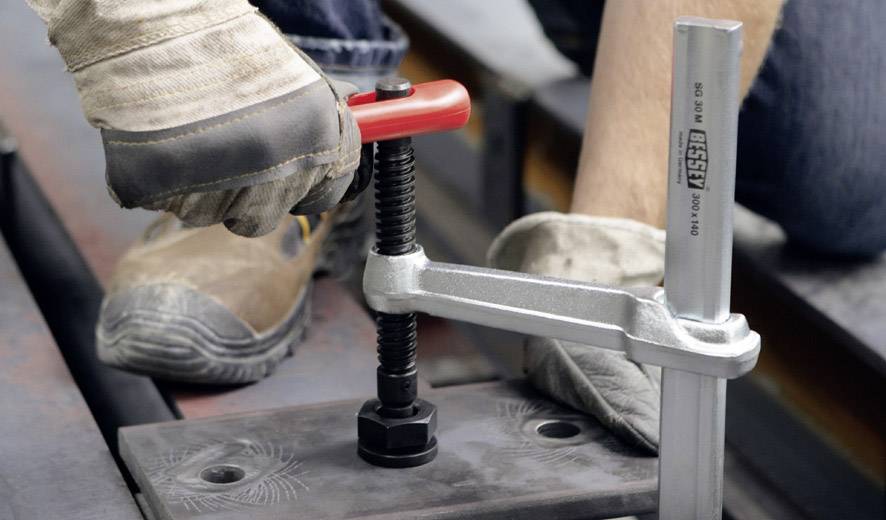 Een persoon met handschoenen gebruikt een rode klembeugel om een werkstuk vast te zetten. De afbeelding toont ambachtelijk werk.