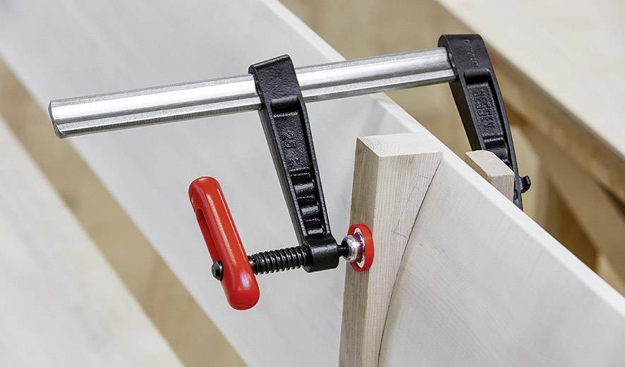 Metalen klem houdt twee houten onderdelen bij elkaar. Een rode schroef dient om vast te draaien. Werkplaatsomgeving zichtbaar op de achtergrond.