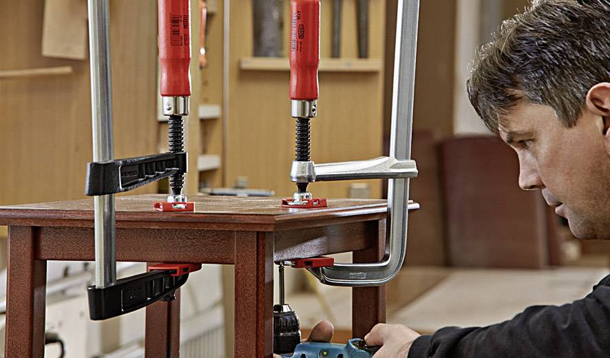 Een ambachtsman bevestigt twee houten onderdelen aan een tafel met klemmen en werkt zorgvuldig eraan met een blauw gereedschap.