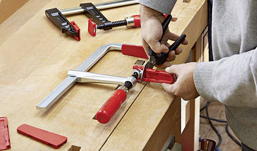 Een persoon gebruikt een bankschroef op een houten tafel. Verspreid liggende rode klemmen en gereedschappen wijzen op een werksituatie.