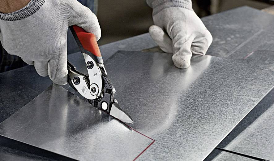 Een persoon knipt een stuk metaal met een metaalschaar. De handen zijn beschermd met grijze handschoenen en werken nauwkeurig.