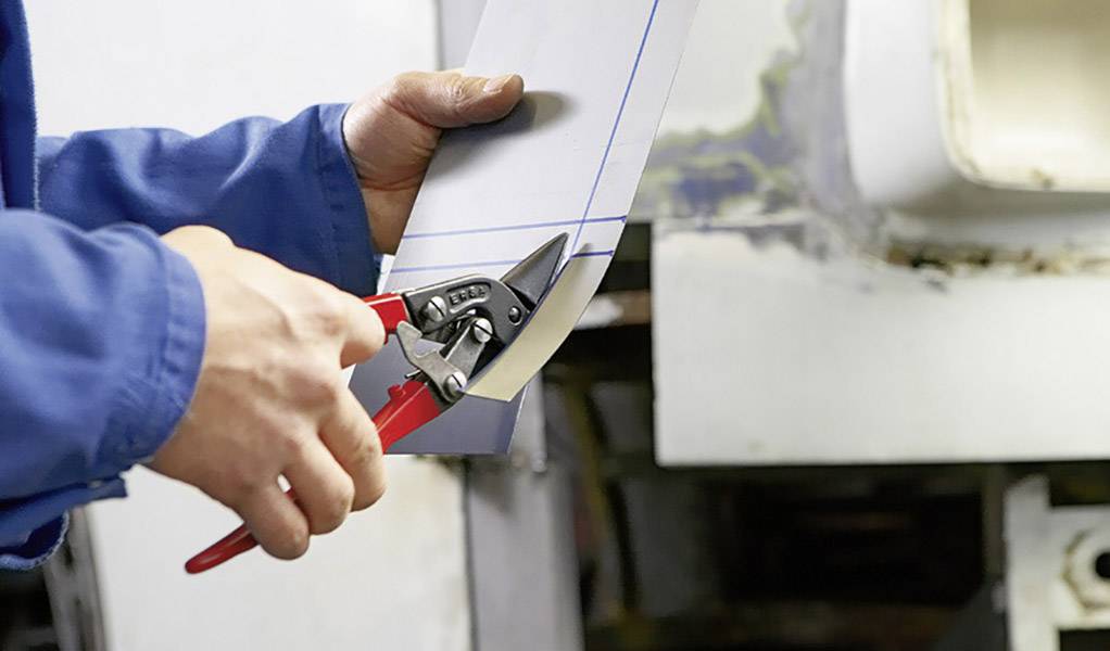 Een persoon in blauwe werkkleding knipt een metalen plaat met een metaalschaar. Op de achtergrond zijn details van een werkplaats zichtbaar.