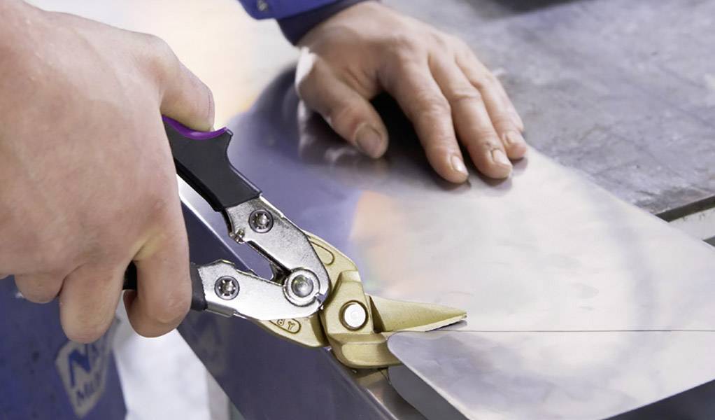 Een persoon knipt een dun metalen plaatje met een metaalschaar. Handen en gereedschap staan centraal in beeld.