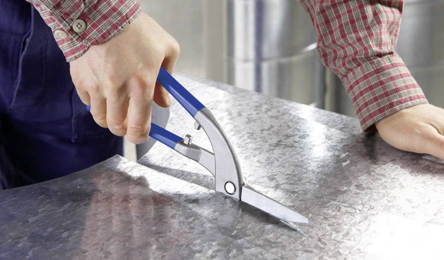 Een persoon knipt een metalen plaat met een blauwe handschaar. De hand draagt een rood geruit overhemd. Scenario: Metaalbewerking.