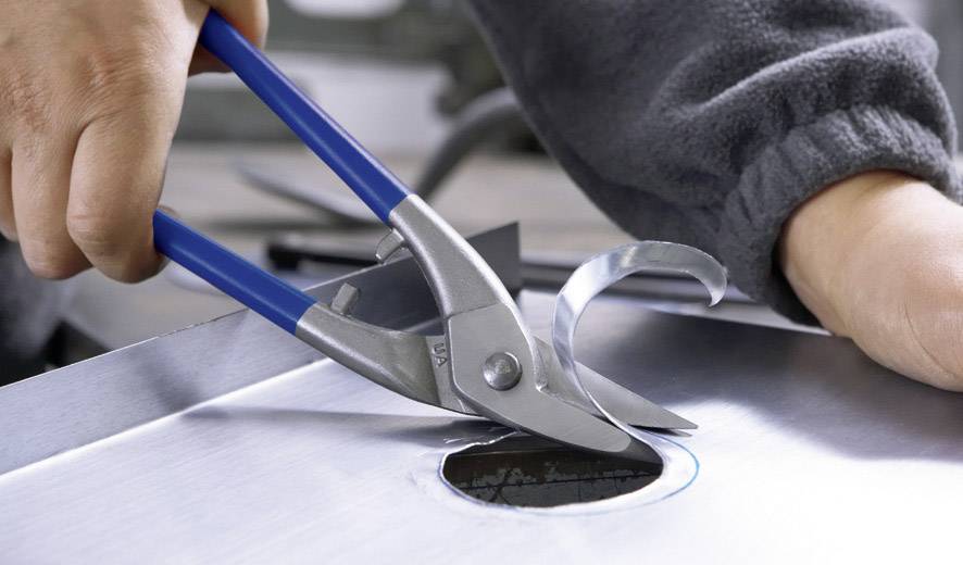 Een persoon knipt met blauwe blikscharen een rond gat in een zilveren metalen plaat.