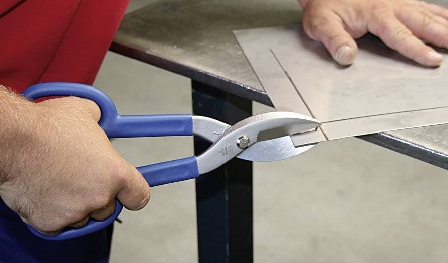 Een persoon knipt een dun metalen plaatje door met een blauwe blikschaar op een tafel.