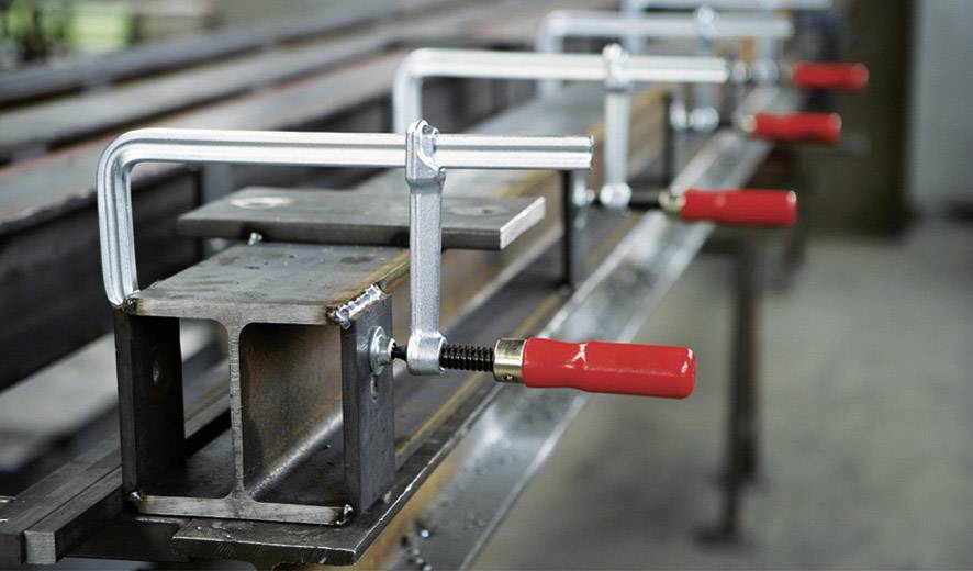 Verschillende metalen klemmen met rode handgrepen houden rechthoekige metalen balken vast op een werkbanktafel.