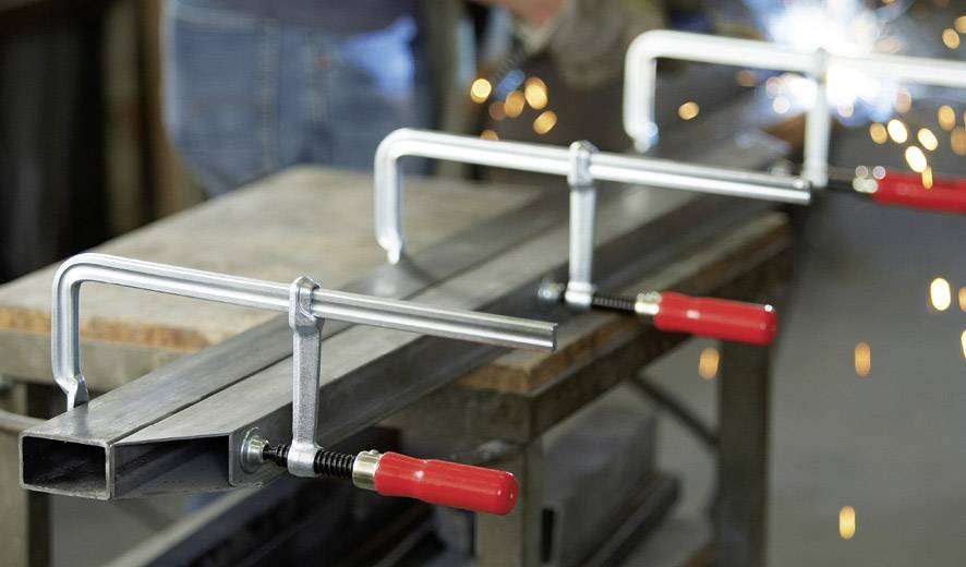 'Close-up van metalen klemmen die een metalen stuk vastzetten op een werkbank. Op de achtergrond zijn vonken van laswerk zichtbaar.'