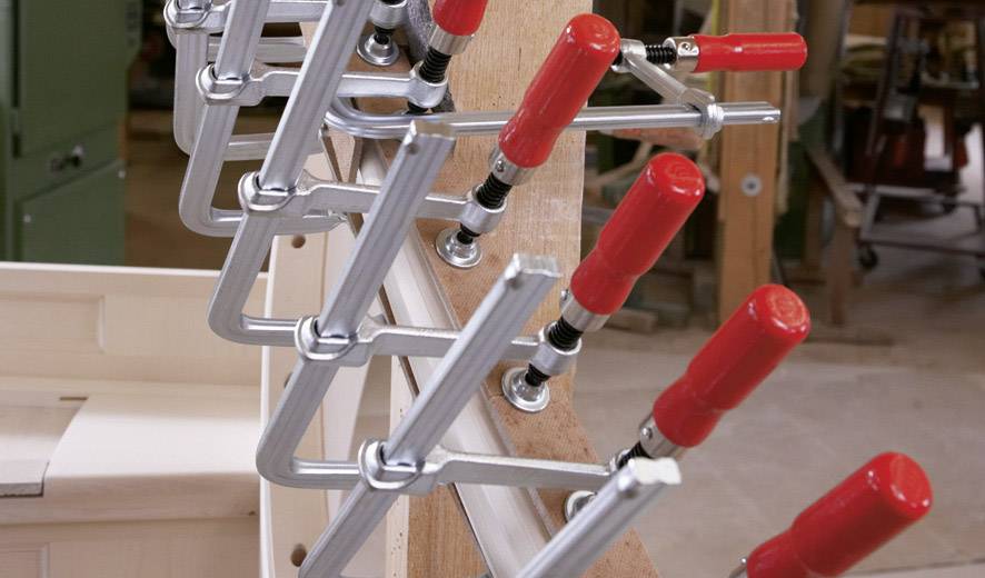 Verschillende lijmklemmen met rode handgrepen houden houten planken stevig vast. Werkomgeving op de achtergrond.