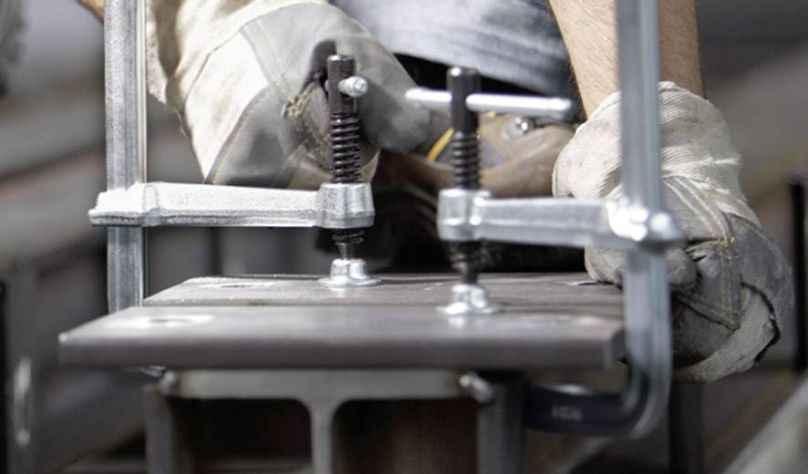 Close-up van een metaalarbeider die een klem gebruikt om twee metalen onderdelen te bevestigen. De arbeider draagt werkhandschoenen.