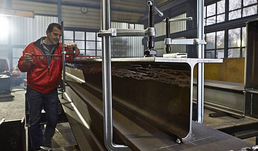 Een man in een rode jas werkt in een werkplaats aan een grote metalen balk, die hij met een gereedschap bewerkt.