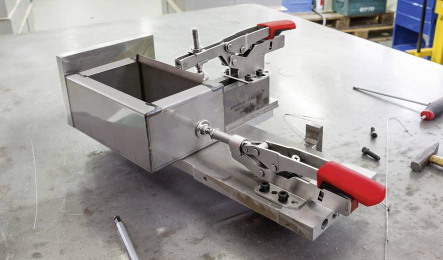 Een metalen apparaat met rode handgrepen staat op een werktafel in een werkplaats. Gereedschappen en schroeven liggen er verspreid omheen.