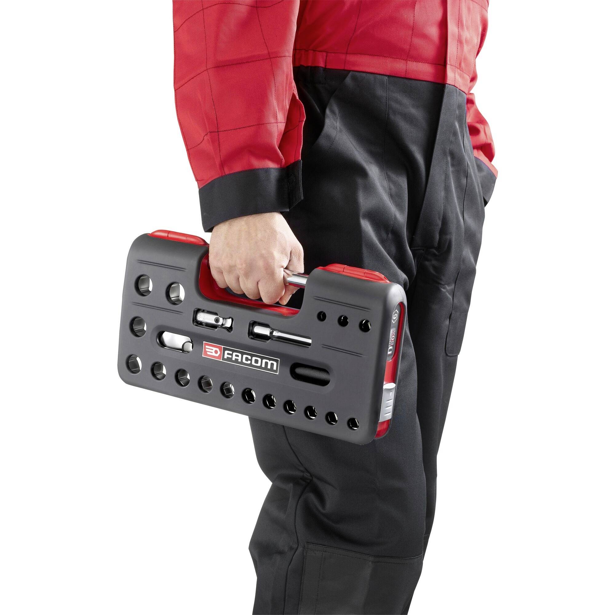 Een persoon houdt een zwarte, geperforeerde, rechthoekige gereedschapskoffer met rode handgreep en het merk 'Facom', gekleed in een rode jas.