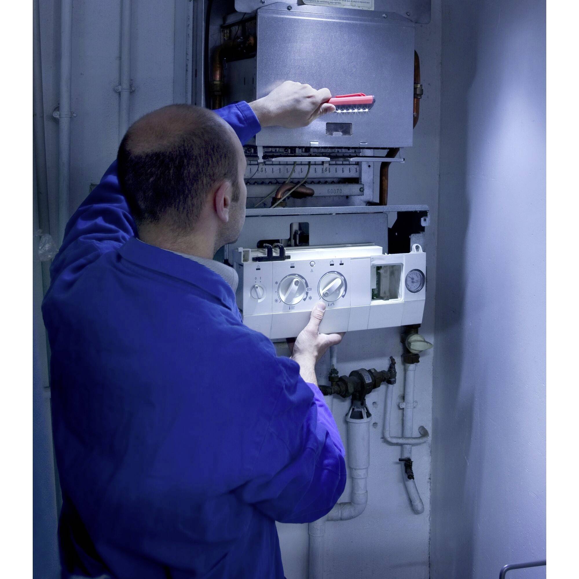 Een technicus in blauwe werkkleding werkt aan een verwarmingsketel. Hij gebruikt een schroevendraaier en controleert de instellingen.